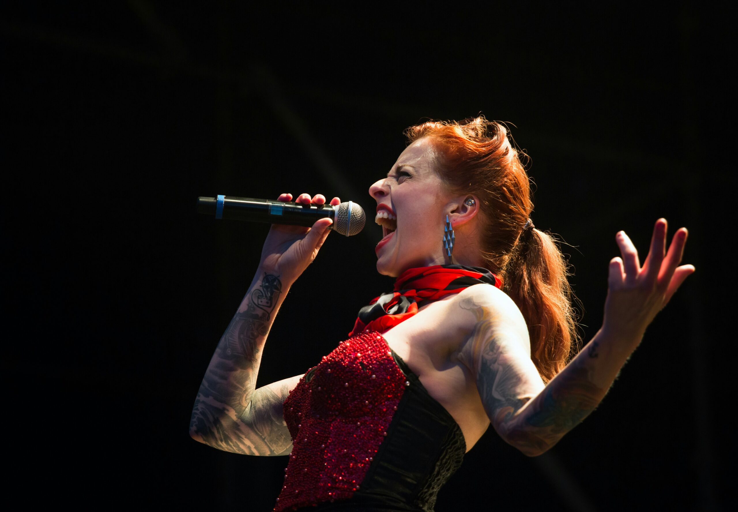 Person with tattooed arms performing on stage with a microphone, wearing a sparkly red and black outfit.