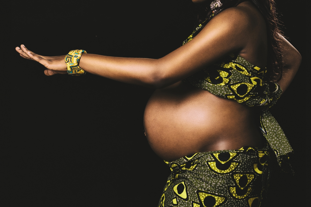 A pregnant person wearing green and yellow patterned clothing, extending their left arm forward against a black background.