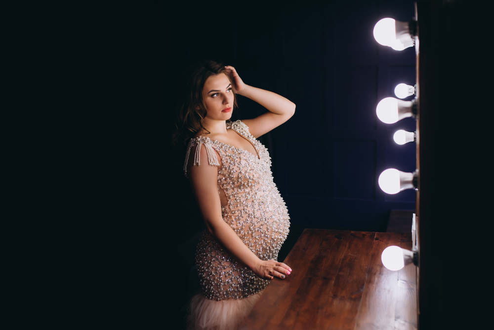 Pregnant woman in a sparkly, embellished dress standing in front of a lit vanity mirror. One hand rests on her belly, the other on her head. The background is dark, with a wooden vanity table visible.