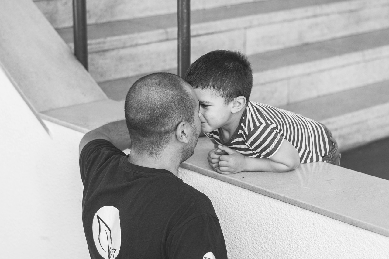 Two individuals interacting near a staircase. One person with a shaved head and a dark shirt featuring a white logo leans against a wall, while a younger person in a striped shirt leans over the same wall from the opposite side. Both faces are obscured.