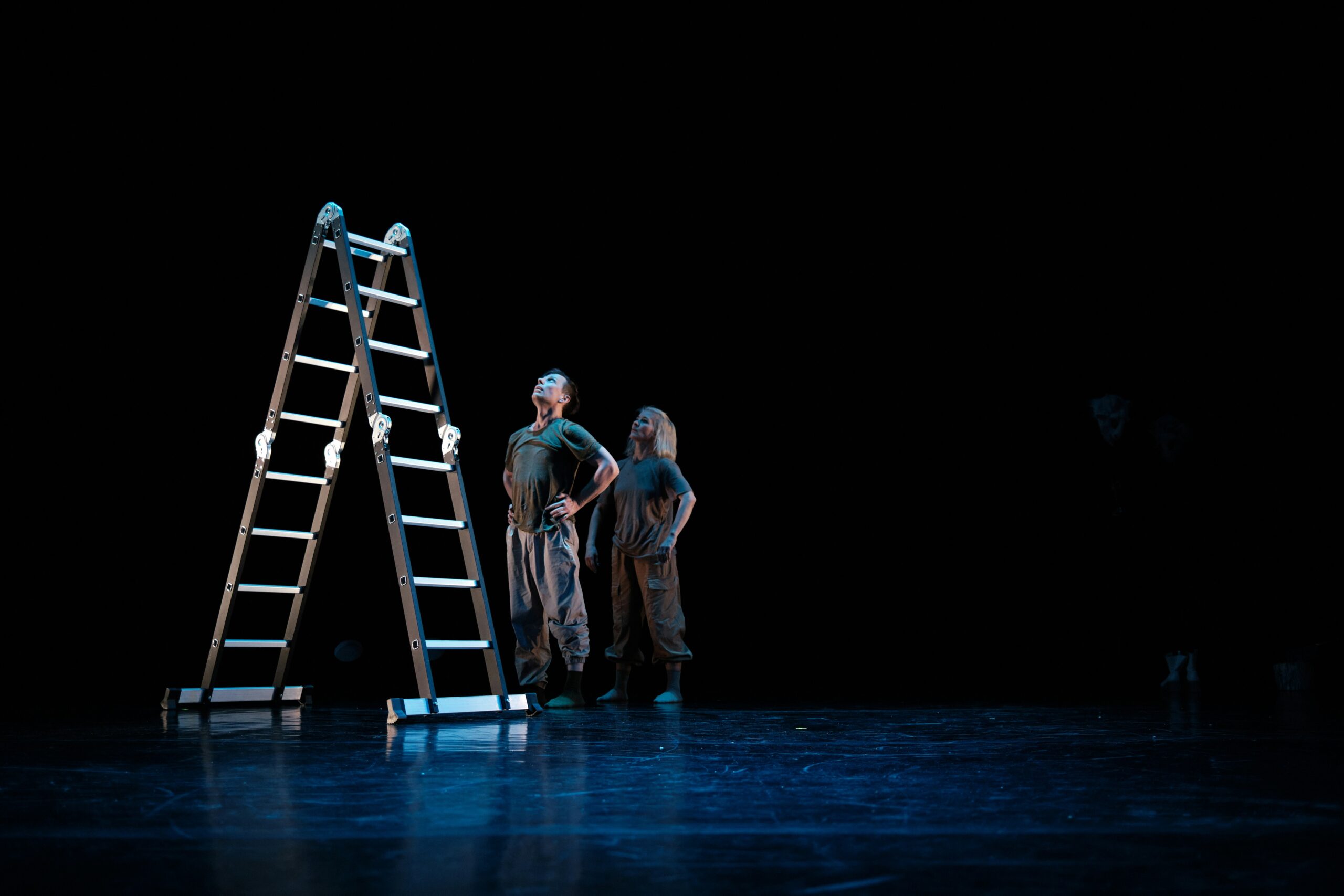 Two dancers wearing uniforms stand beside a tall aluminum ladder on a dimly lit stage, conveying a sense of anticipation.