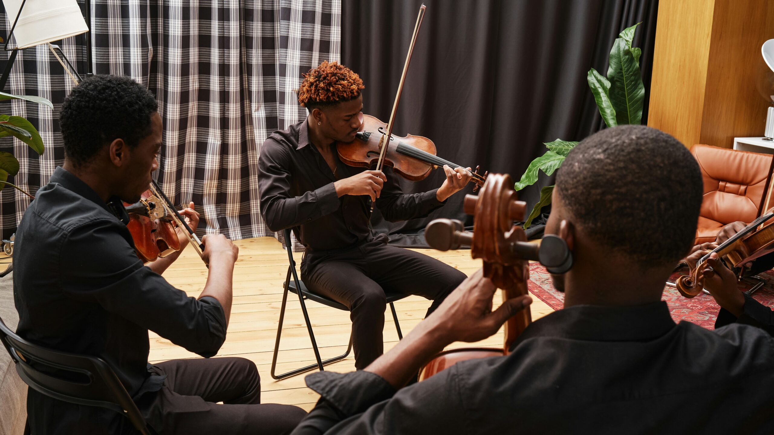 Four individuals seated in a circle playing violins and a viola in a rehearsal setting, with music stands, sheet music, a checkered curtain, and plants in the background.
