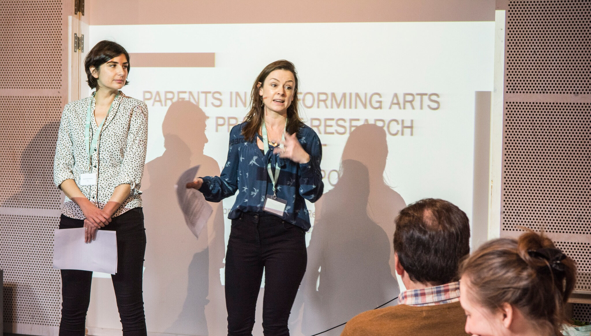 Cassie and Anna presenting at an event in front of an audience, with a projected slide in the background.