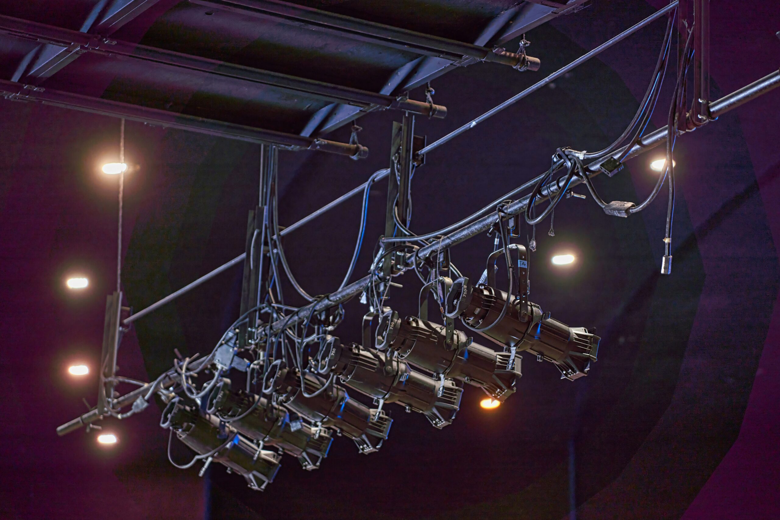 Stage lights mounted on a metal rigging system, illuminated against a dark background with visible connecting cables.