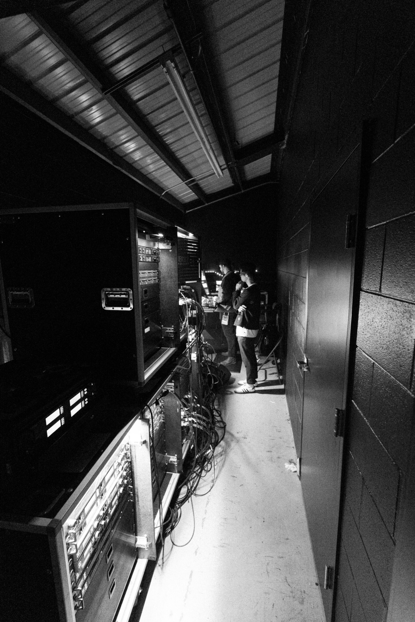 Black and white photo of a dimly lit backstage area with people working on electronic equipment, surrounded by cables, racks, and exposed ceiling beams.