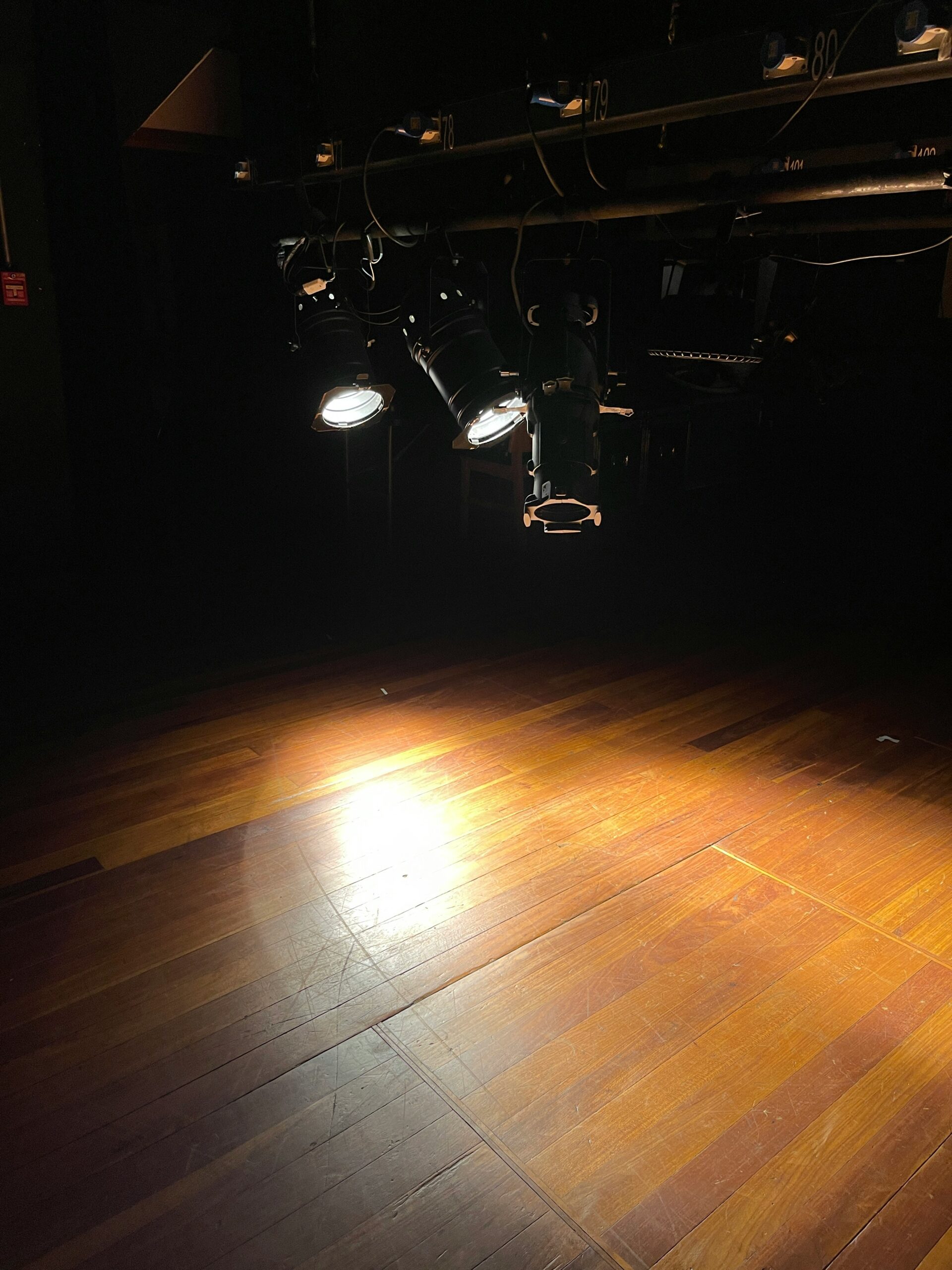 A stage with wooden flooring lit by overhead stage lights, creating a bright spotlight in an otherwise dark setting.