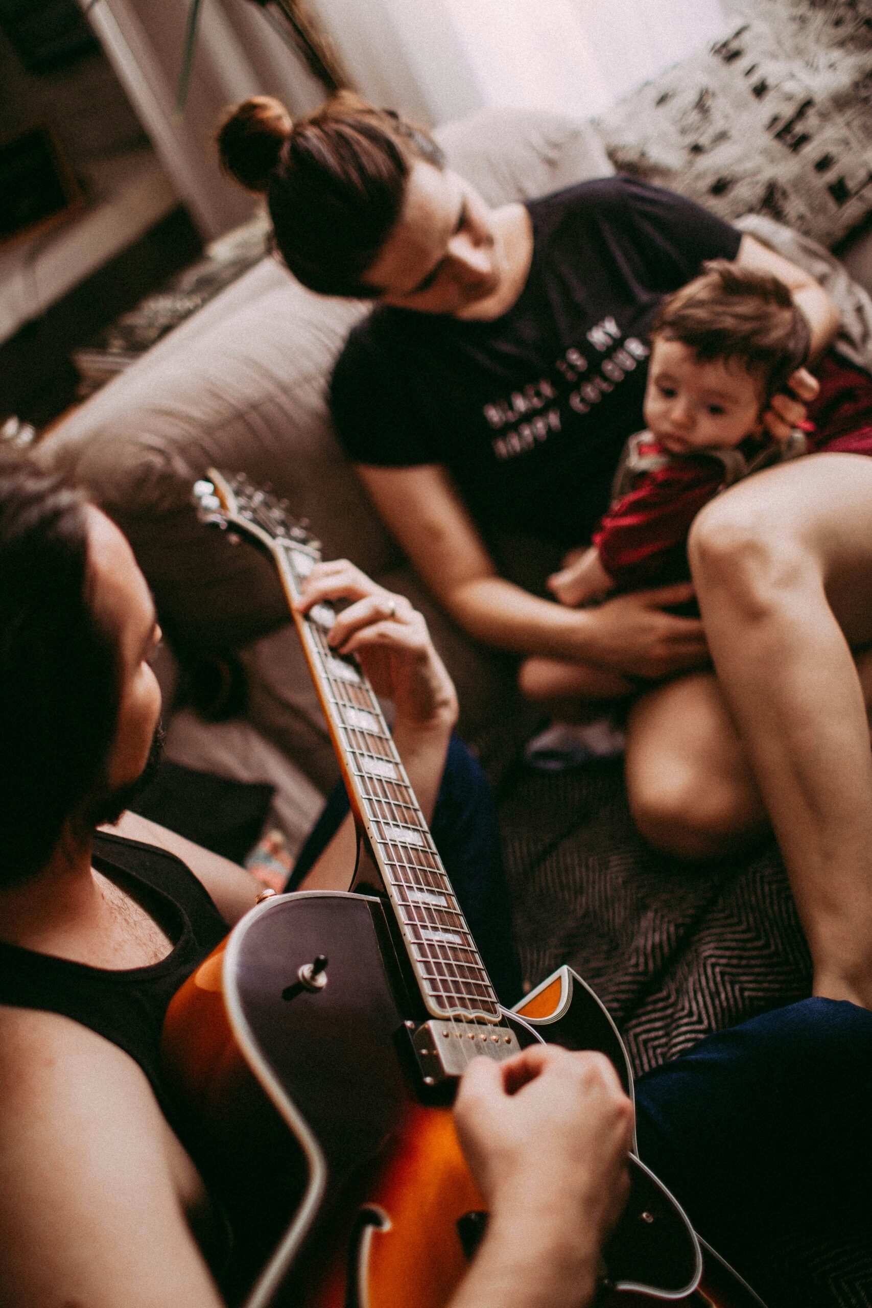 Three people in a cozy indoor setting, one playing an electric guitar and another holding a child on their lap, with faces blurred for privacy. The person holding the child wears a shirt that says 'MAKE ART NOT WAR.