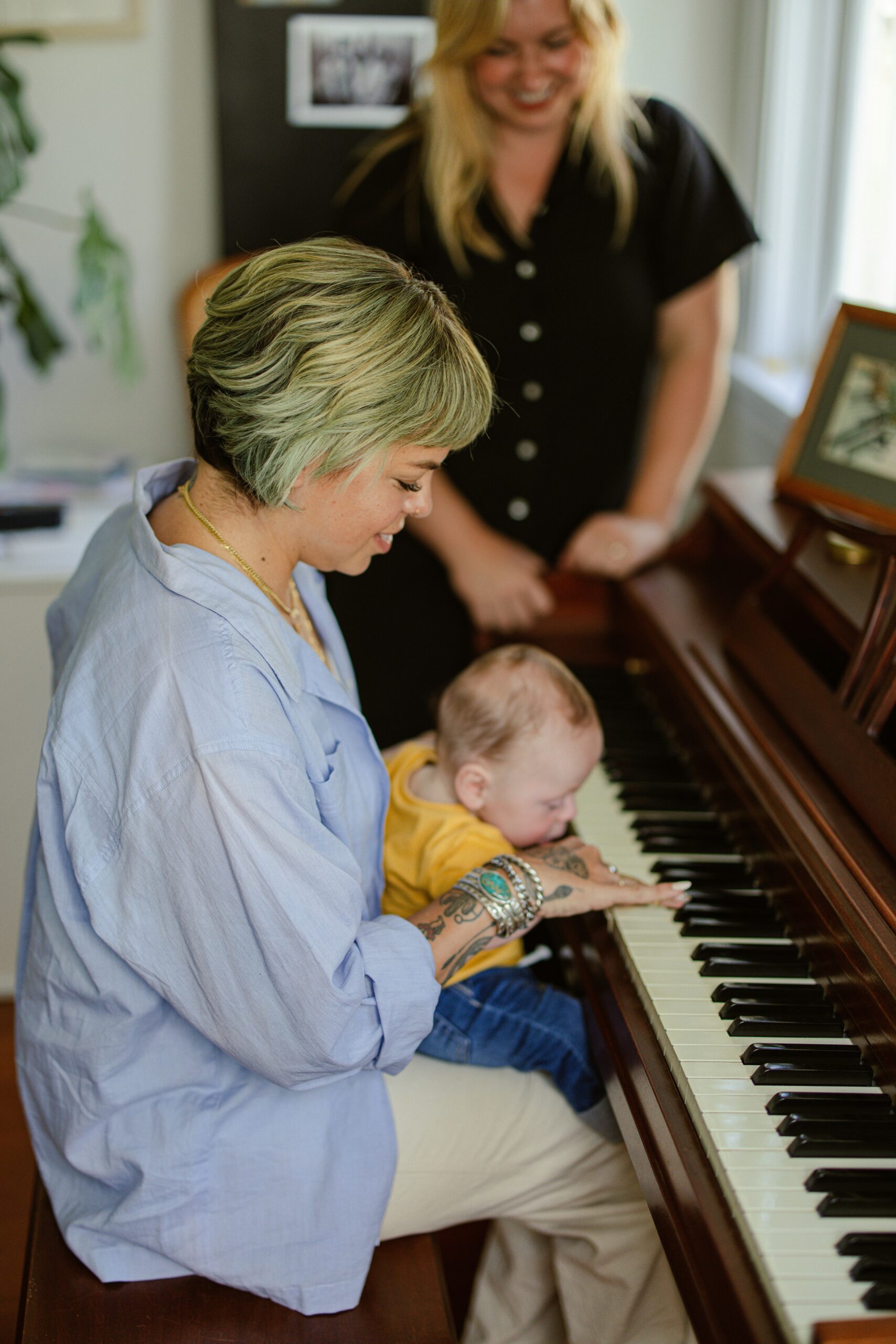 Two adults and a baby at a piano. One adult is seated, holding the baby on their lap and guiding the baby's hands on the piano keys, while the other adult stands behind them. A window and framed pictures are visible in the background