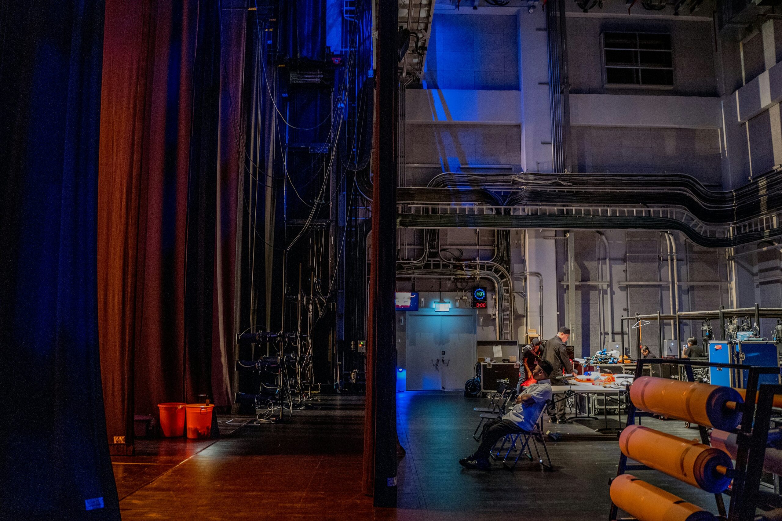 Backstage area of a theater with stage curtains and lighting equipment on the left, a partition wall in the center, and people working at tables with technical gear on the right under blue lighting.