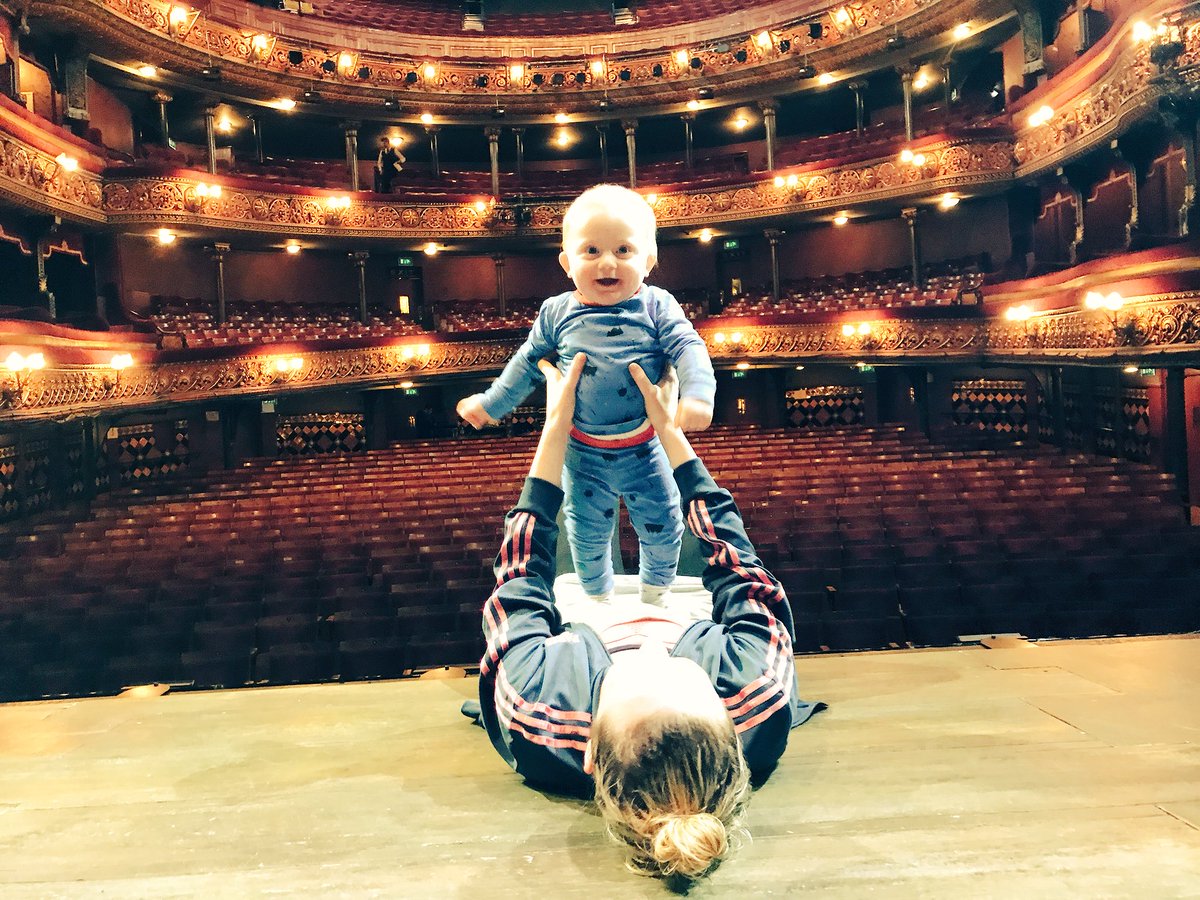 Person lying on a stage holding a small child aloft with their hands, set against the backdrop of an empty theater with ornate balconies and seating