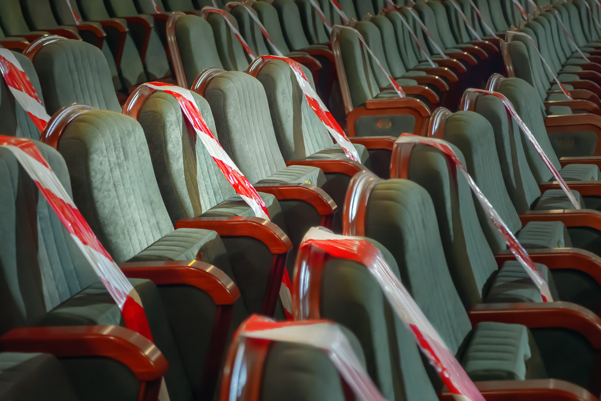 Rows of green upholstered theater seats with wooden armrests, some marked with red and white caution tape to indicate restricted use.