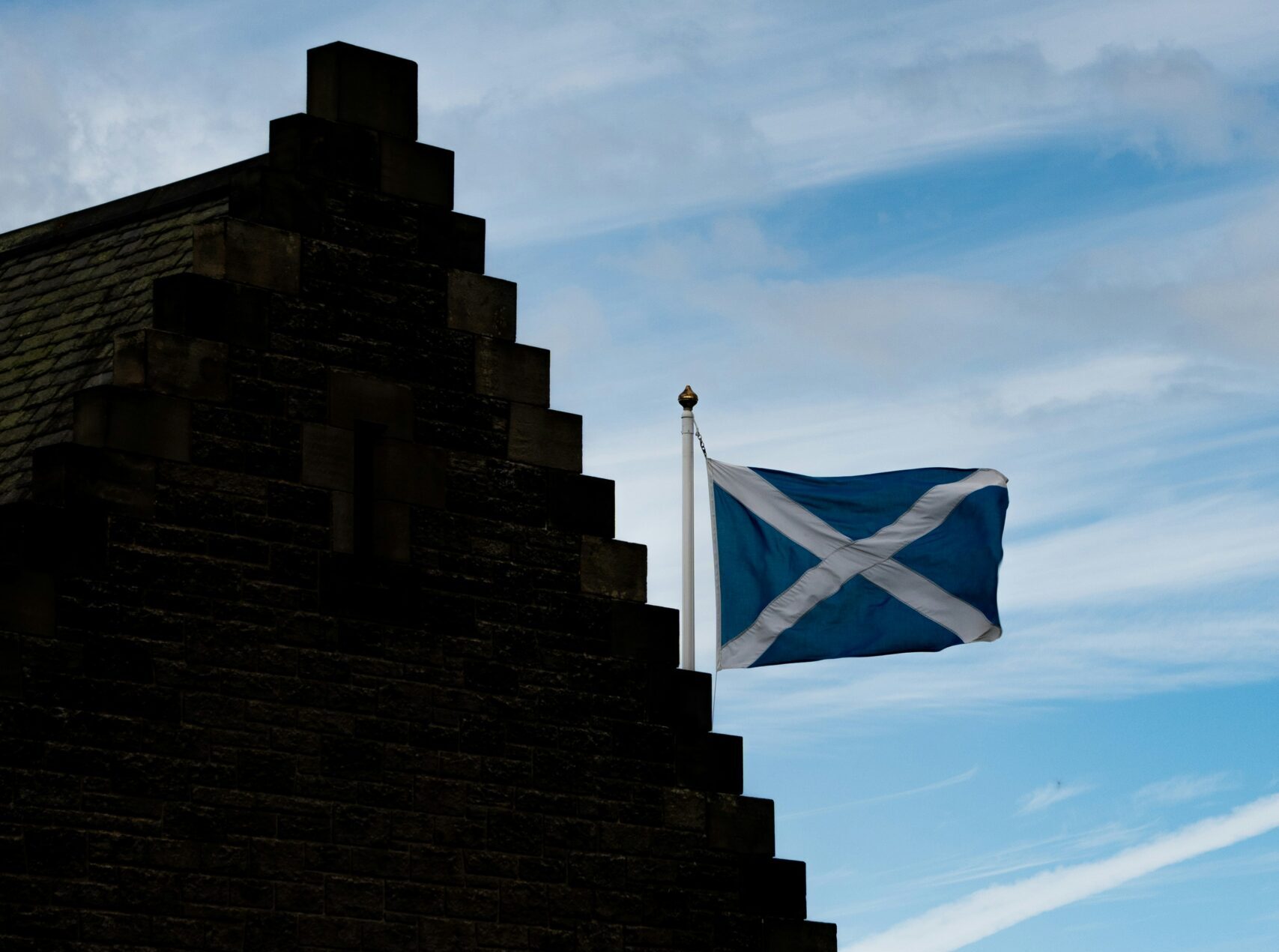 A building with a stepped gable roof and the Scottish flag flying on a flagpole, set against a partly cloudy sky with patches of blue.