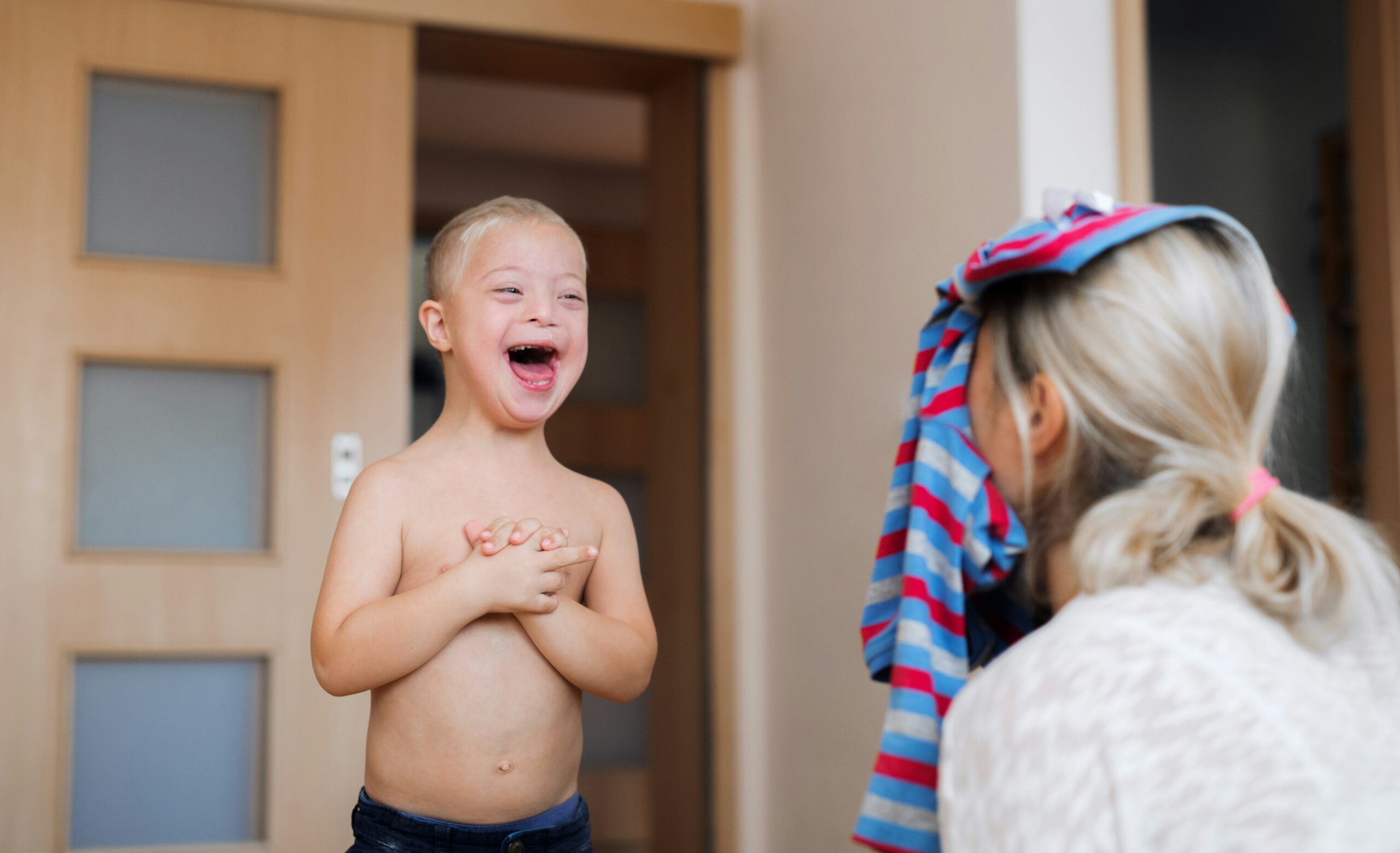 A child stands with hands clasped, wearing shorts, while an adult with long hair faces them, they have a long sleeve t shirt over their face.